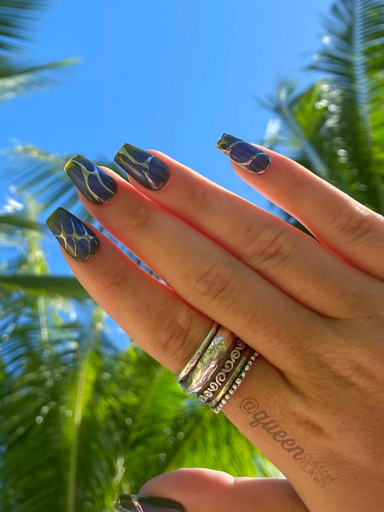 hand held up, modeling custom made press on nails with an aura heart, and 90's tribal chrome designs in silver. each nail has a different design in chrome. the nail base is purple, with an airbrushed center in a darker blue over the purple base. the other hand is olive green and purple base colors under the silver designs