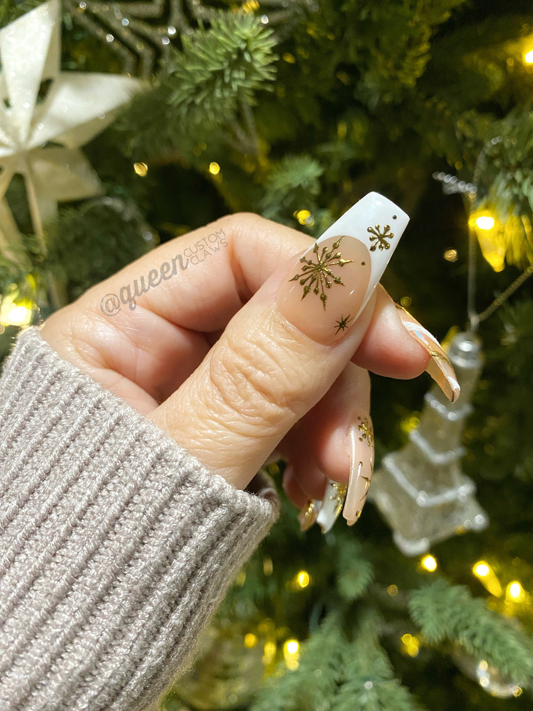 a person showing their Christmas themed press on nails in front of a christmas tree. The press on nails are pink and white French tips with gold snowflakes hand painted with gold chrome